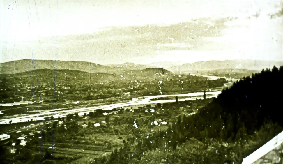 Peisaj panoramic ce prezintă o zonă de munte cu un râu care curge printr-o vale, cu case și câmpuri vizibile în depărtare.