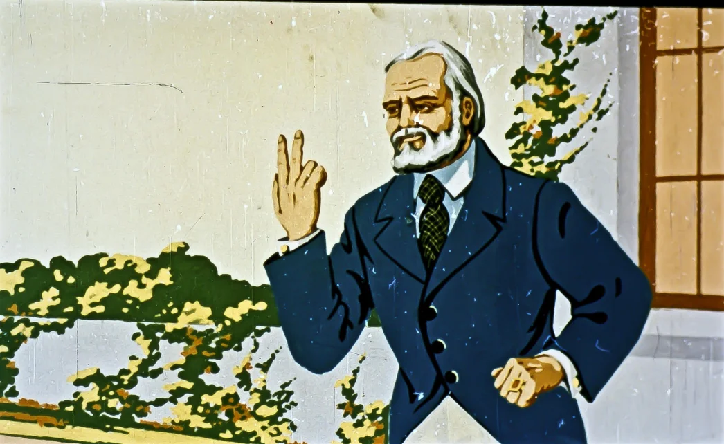 Imaginea unui bărbat cu părul și barba albă, îmbrăcat într-un costum albastru, care ridică degetul arătător, pe fondul unei pereți cu feronerie și plante decorative.